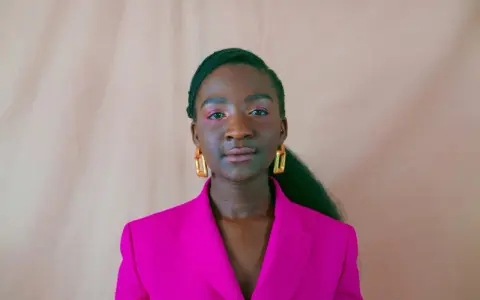 Een jonge vrouw van kleur met opvallende gouden oorbellen en een fuchsia blazer, kijkt zelfverzekerd de camera in.