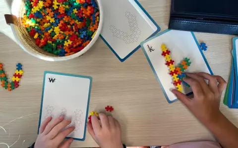 Kinderen bouwen met kleurrijke stukjes aan patronen op kaarten. Een mand vol bouwstenen ligt klaar voor meer taal spelletjes.