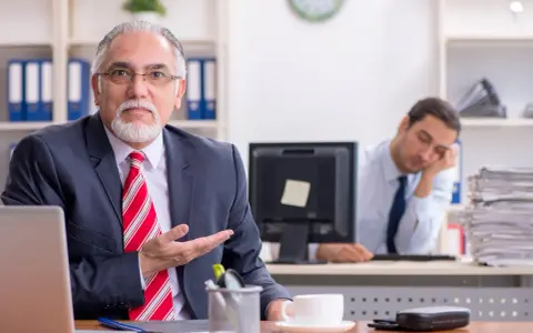 Een oudere man in pak legt iets uit, terwijl een jongere collega achter hem in slaap valt. Een beeld van volwassen worden en de uitdagingen van het werk.