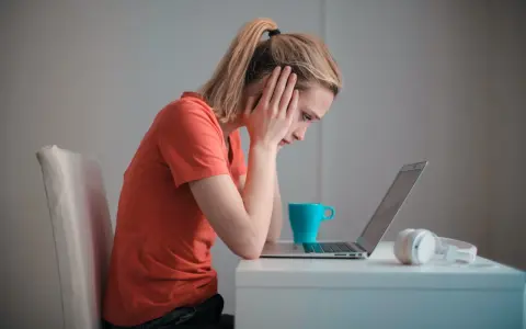 Een meisje met een korte spanningsboog staart naar haar laptop, handen aan haar hoofd. Een blauwe mok en koptelefoon staan naast haar.