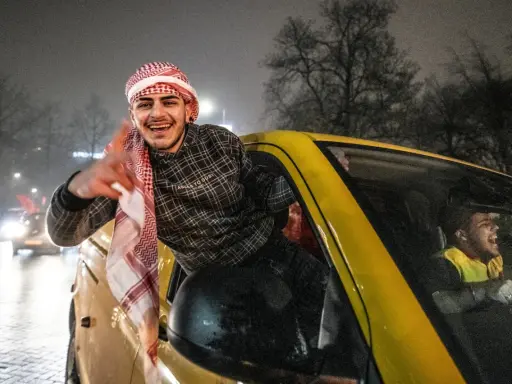 Jonge Marokkaanse Nederlanders vieren feest in een gele auto, met een van hen die uit het raam hangt en lachend zwaait.