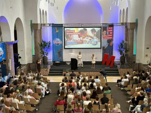 Publiek luistert naar presentatie over "de wereld in getallen". Twee sprekers op podium, met projectiescherm en grote rode M.