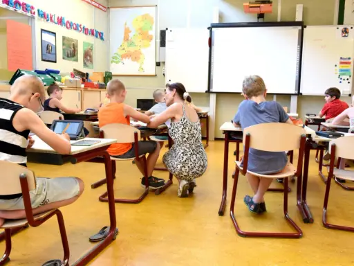 Een klas met kinderen die geconcentreerd aan hun tafels zitten. Een juf helpt een leerling, terwijl anderen werken op tablets of met potloden.