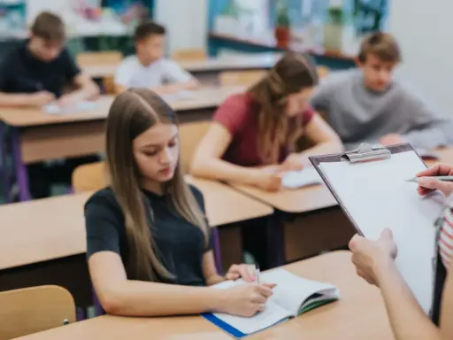 Leerlingen in een klaslokaal, sommigen schrijven in hun schrift. De leraar houdt een clipboard vast, misschien om te zien wat zijn leerlingen leren over wat zijn beta vakken.