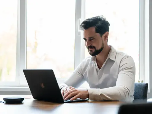 Man met baard, gekleed in een wit overhemd, geconcentreerd op zijn werk achter een laptop.