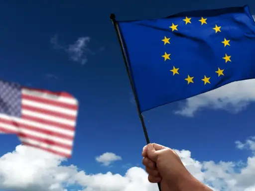 Hand holds EU flag, US flag blurred in background. Discussing what is soevereiniteit in a global context.