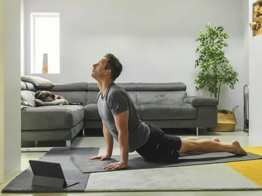 Man in cobra pose op yogamat, neemt regie over zijn leven. Hond slaapt op bank.