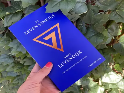 Een hand houdt het boek "De Zeven Vinkjes" van Joris Luyendijk vast, omringd door groen blad.