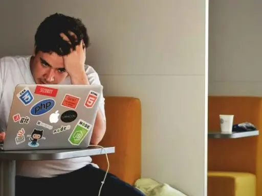 Een gefrustreerde jongere met zijn hand in zijn haar, starend naar zijn laptop vol met programmeerstickers. De druk van jongeren en schulden kan voelen als een eindeloze code.