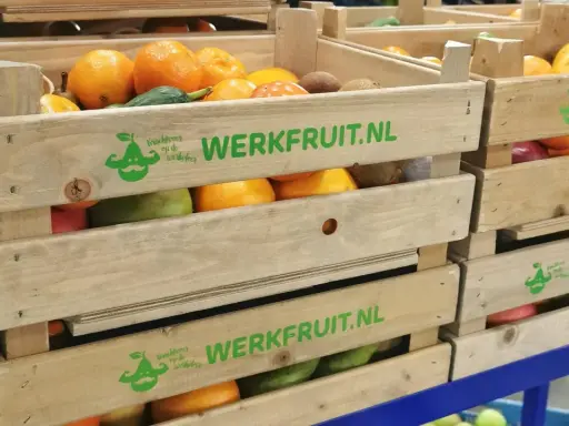 Houten kratten met fruit, waaronder sinaasappels en kiwi's, met de tekst "WERKFRUIT.NL" en een logo dat staat voor duurzame inzetbaarheid.
