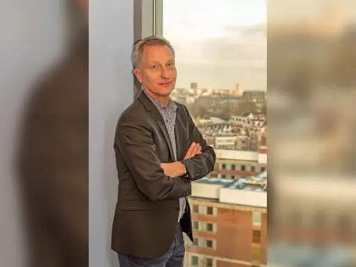 Jan Willem Duyvendak, een man met een grijze blazer en een geruit overhemd, staat met gekruiste armen voor een raam met uitzicht op een stad.