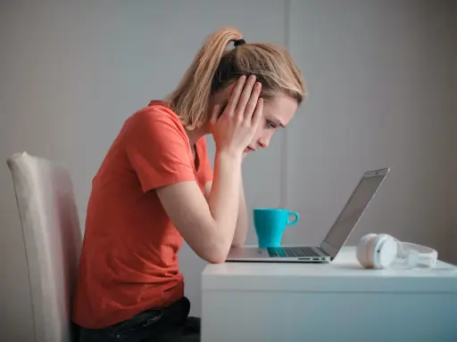 Een meisje met een korte spanningsboog staart naar haar laptop, handen aan haar hoofd. Een blauwe mok en koptelefoon staan naast haar.