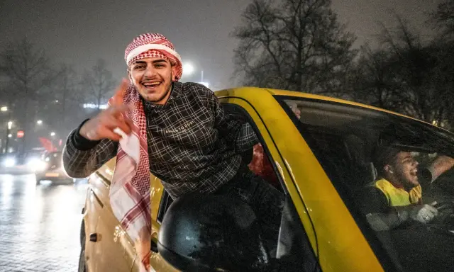Jonge Marokkaanse Nederlanders vieren feest in een gele auto, met een van hen die uit het raam hangt en lachend zwaait.
