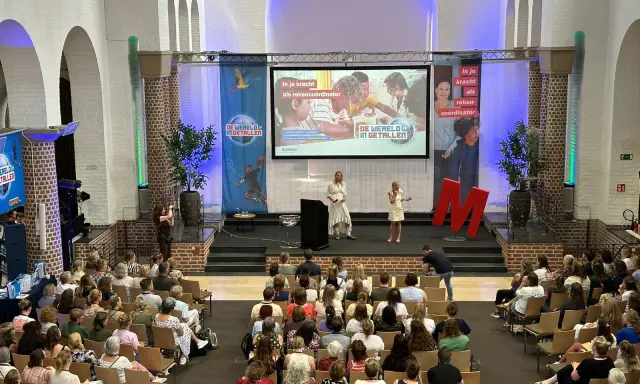 Publiek luistert naar presentatie over "de wereld in getallen". Twee sprekers op podium, met projectiescherm en grote rode M.