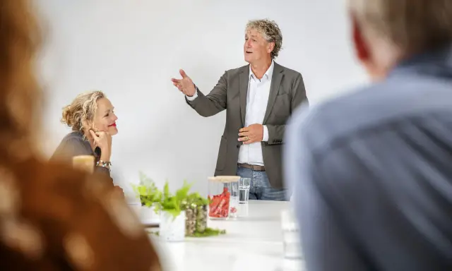 Een man spreekt enthousiast in een vergaderruimte, terwijl een vrouw aandachtig luistert. Ze vormen samen een sterk team.
