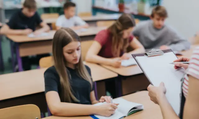 Leerlingen in een klaslokaal, sommigen schrijven in hun schrift. De leraar houdt een clipboard vast, misschien om te zien wat zijn leerlingen leren over wat zijn beta vakken.