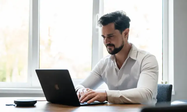 Man met baard, gekleed in een wit overhemd, geconcentreerd op zijn werk achter een laptop.