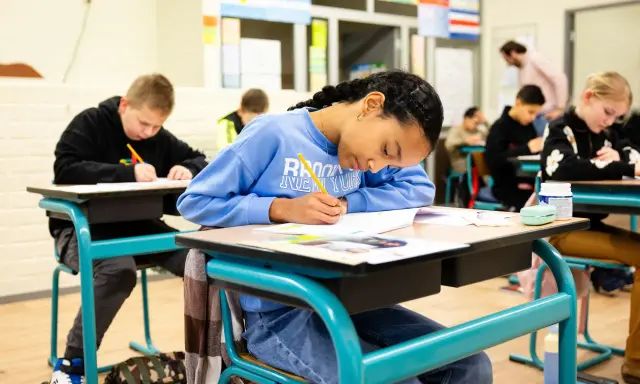 Kinderen maken hun werk, waarbij het leren zichtbaar wordt op papier. Een meisje met een blauwe trui schrijft geconcentreerd.
