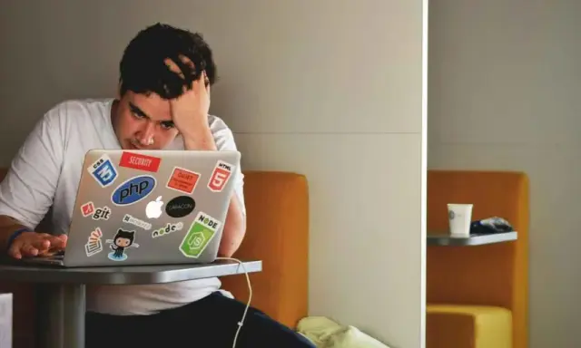Een gefrustreerde jongere met zijn hand in zijn haar, starend naar zijn laptop vol met programmeerstickers. De druk van jongeren en schulden kan voelen als een eindeloze code.