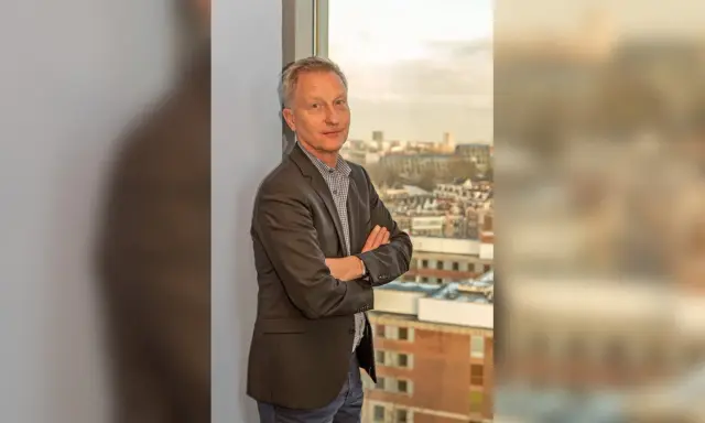 Jan Willem Duyvendak, een man met een grijze blazer en een geruit overhemd, staat met gekruiste armen voor een raam met uitzicht op een stad.