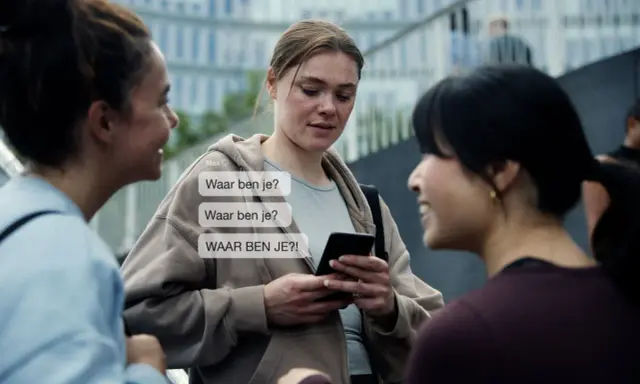 Drie vrouwen praten, één kijkt bezorgd naar haar telefoon met berichten als "Waar ben je?". Dit kan duiden op de angst die gepaard gaat met huiselijk geweld bij vrouwen.