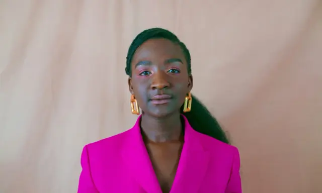 Een jonge vrouw van kleur met opvallende gouden oorbellen en een fuchsia blazer, kijkt zelfverzekerd de camera in.