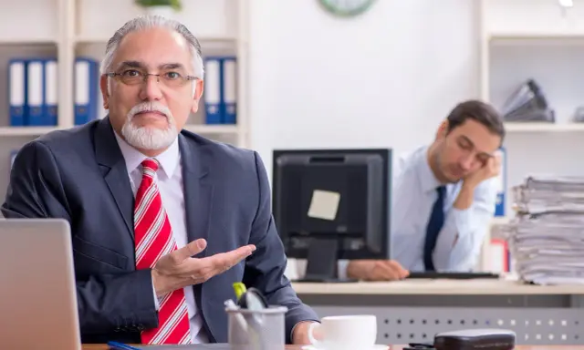 Een oudere man in pak legt iets uit, terwijl een jongere collega achter hem in slaap valt. Een beeld van volwassen worden en de uitdagingen van het werk.