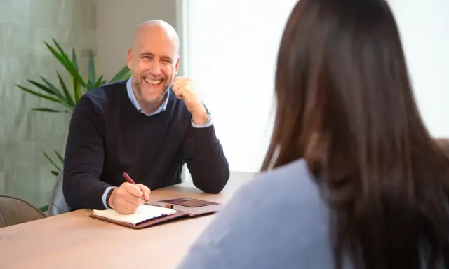 Man met baard en blauwe trui lacht en voert een beoordelingsgesprek, schrijft aantekeningen in een notitieboekje.