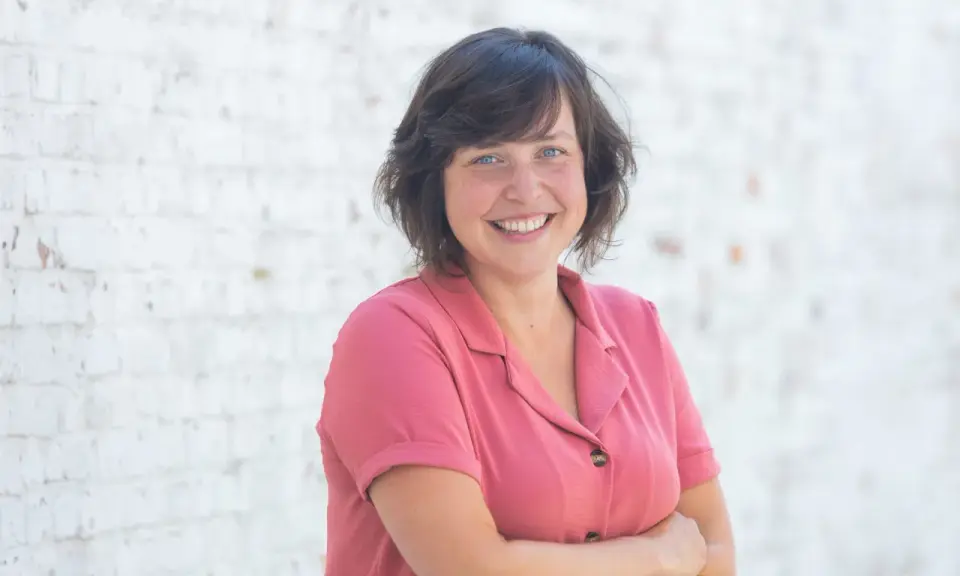 Een lachende vrouw met blauwe ogen, gekleed in een roze blouse, staat voor een witte bakstenen muur. Ze straalt de warmte uit van een zorgzame buurt.