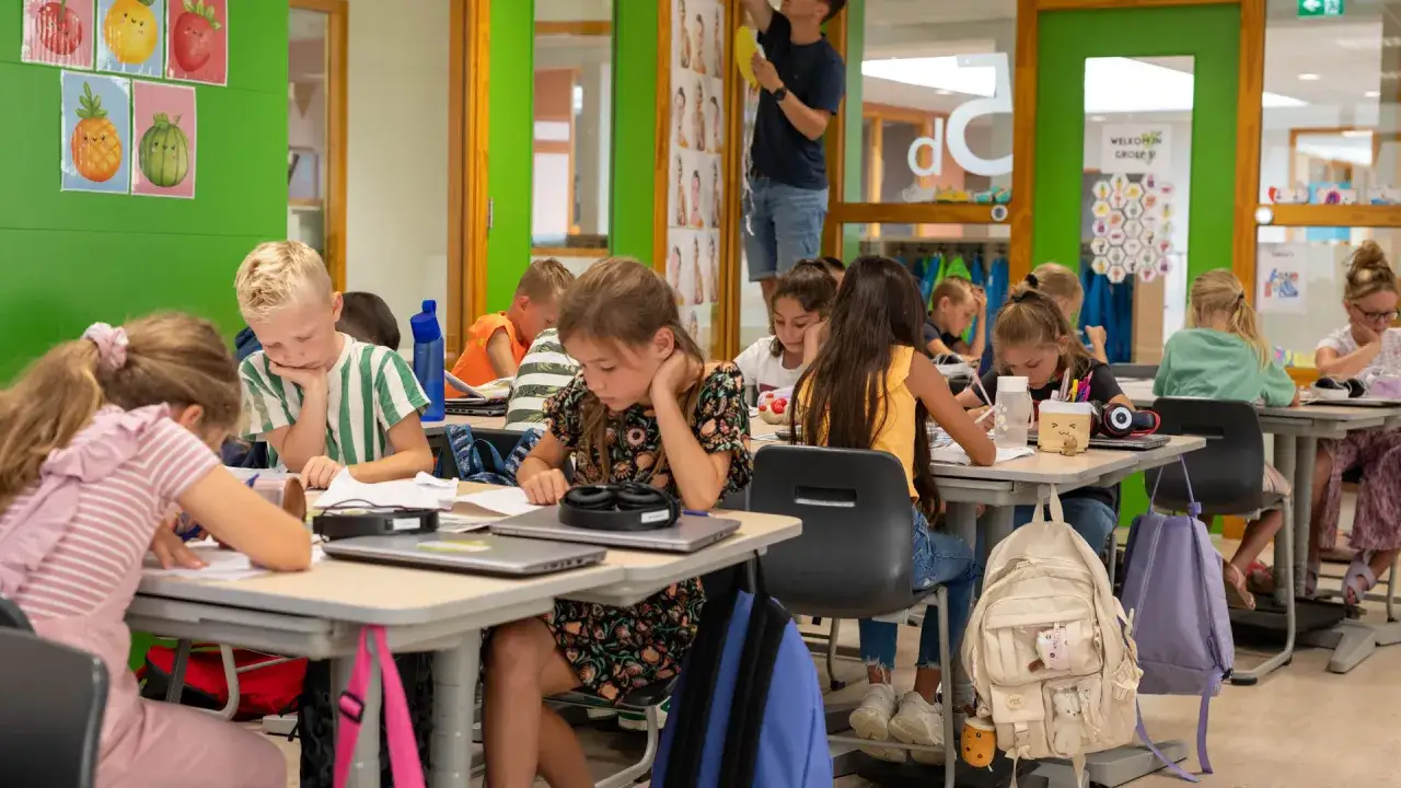 Welkom in groep 5! Kinderen werken geconcentreerd aan hun tafels, omringd door vrolijke fruitposters en schoolspullen.
