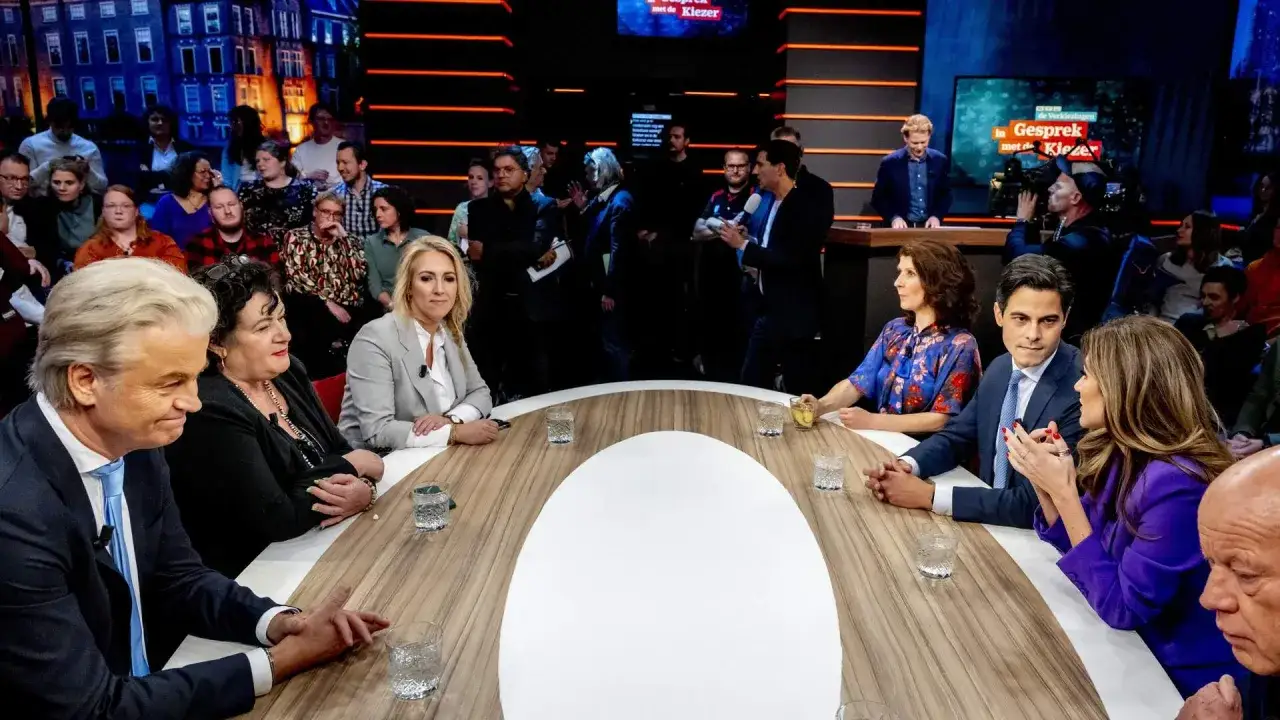 Een groep mensen zit aan een ronde tafel, discussiërend over beleid en maatschappij. De sfeer is gespannen, met serieuze gezichten en geconcentreerde blikken.