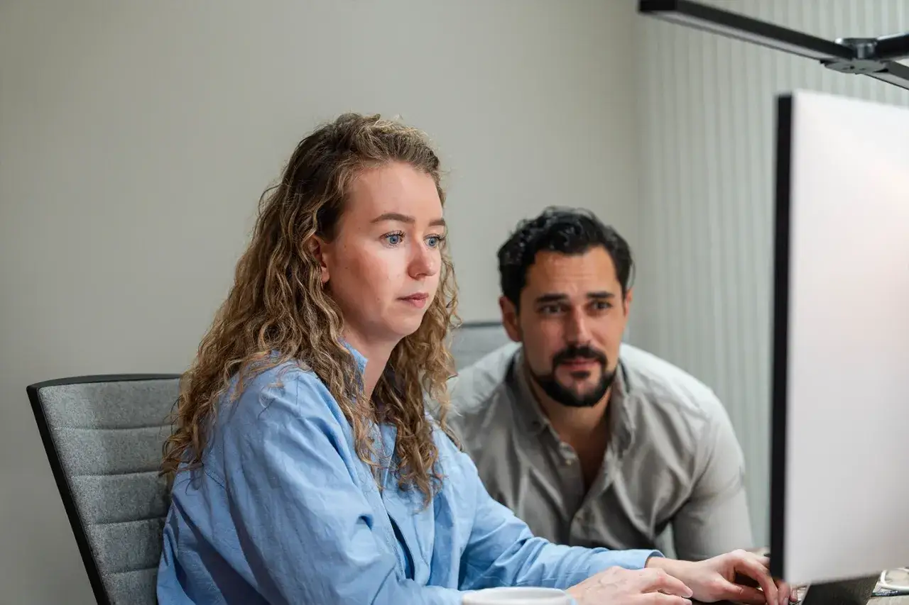 Vrouw met krullend haar en man kijken geconcentreerd naar een computerscherm. Dit toont sturend leiderschap in een team.