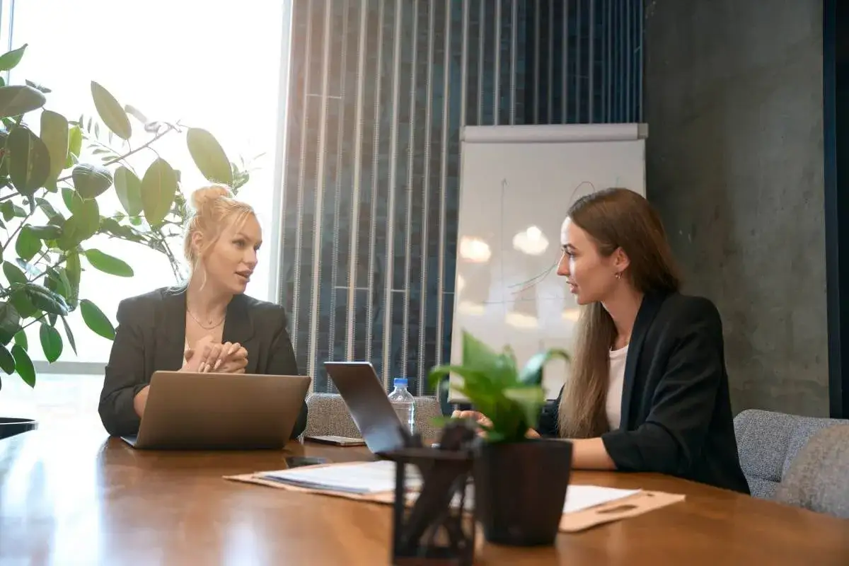 Twee zakenvrouwen bespreken argumenten voor salarisverhoging aan een houten tafel, met laptops en een flip-over op de achtergrond.