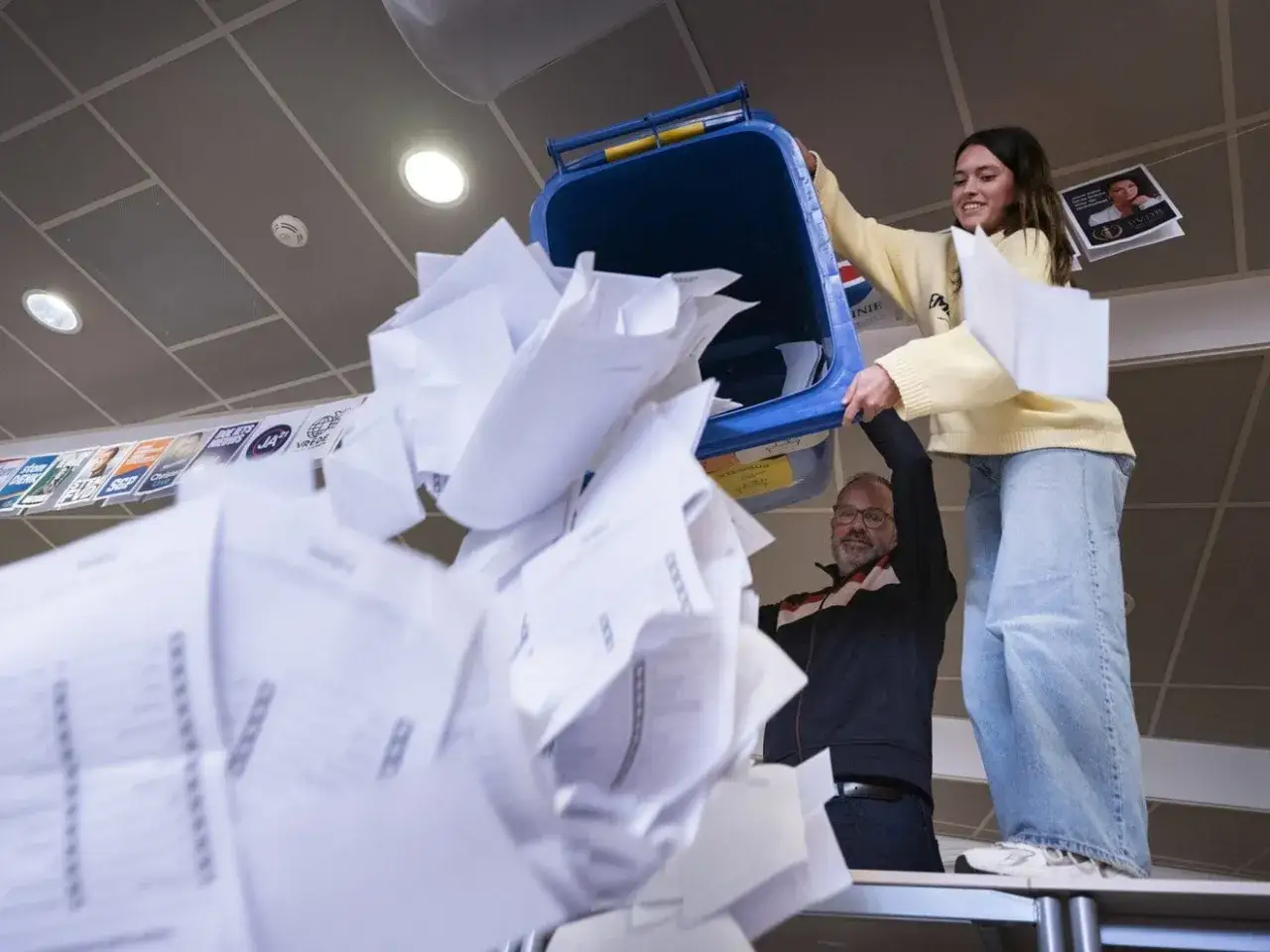 Een jongedame en een man gooien papier in een blauwe bak. Op de achtergrond hangen verkiezingsposters, waaronder een van de PVV.