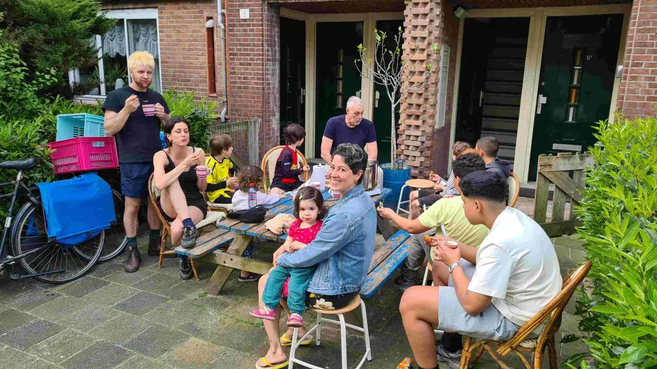 Een groep mensen geniet van een gezellige picknick in de tuin. Kinderen en volwassenen zitten aan een picknicktafel, wat de sociale cohesie en het samenzijn bevorderen.