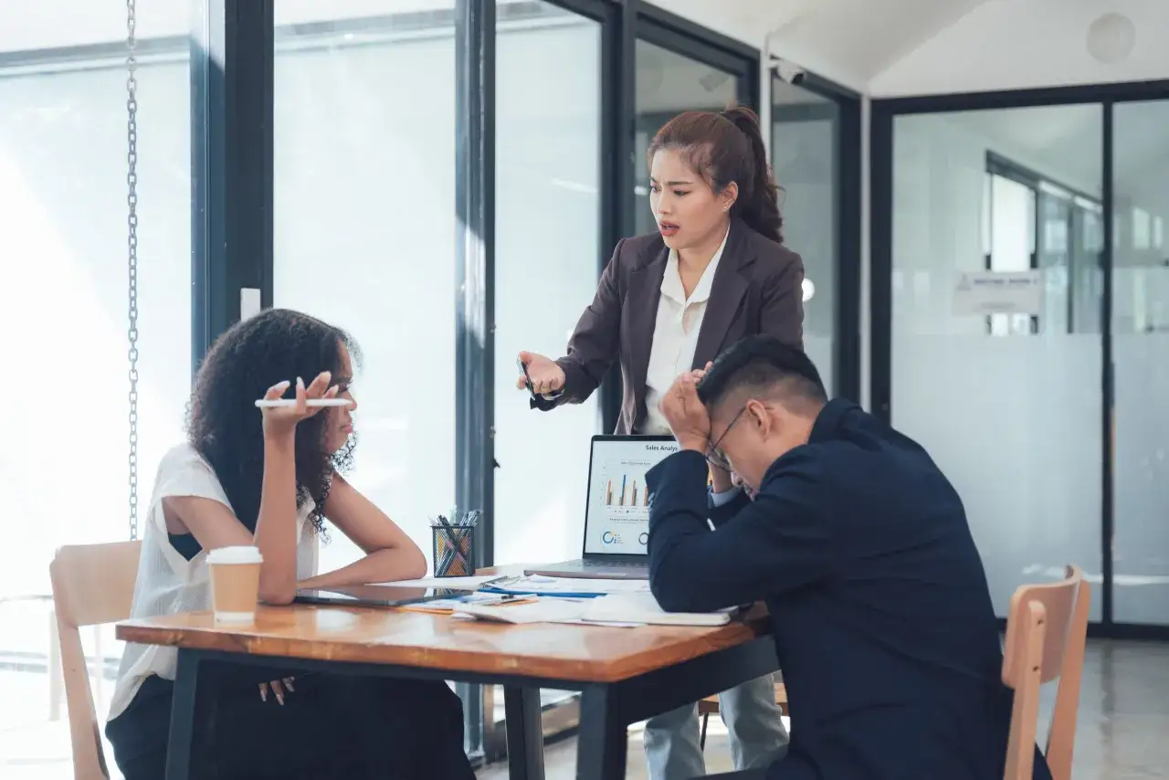 Drie collega's in een vergadering, met een vrouw die gefrustreerd praat, een man met zijn hoofd in zijn handen en een vrouw die luistert. Dit toont de uitdagingen van interpersoonlijke intelligentie.
