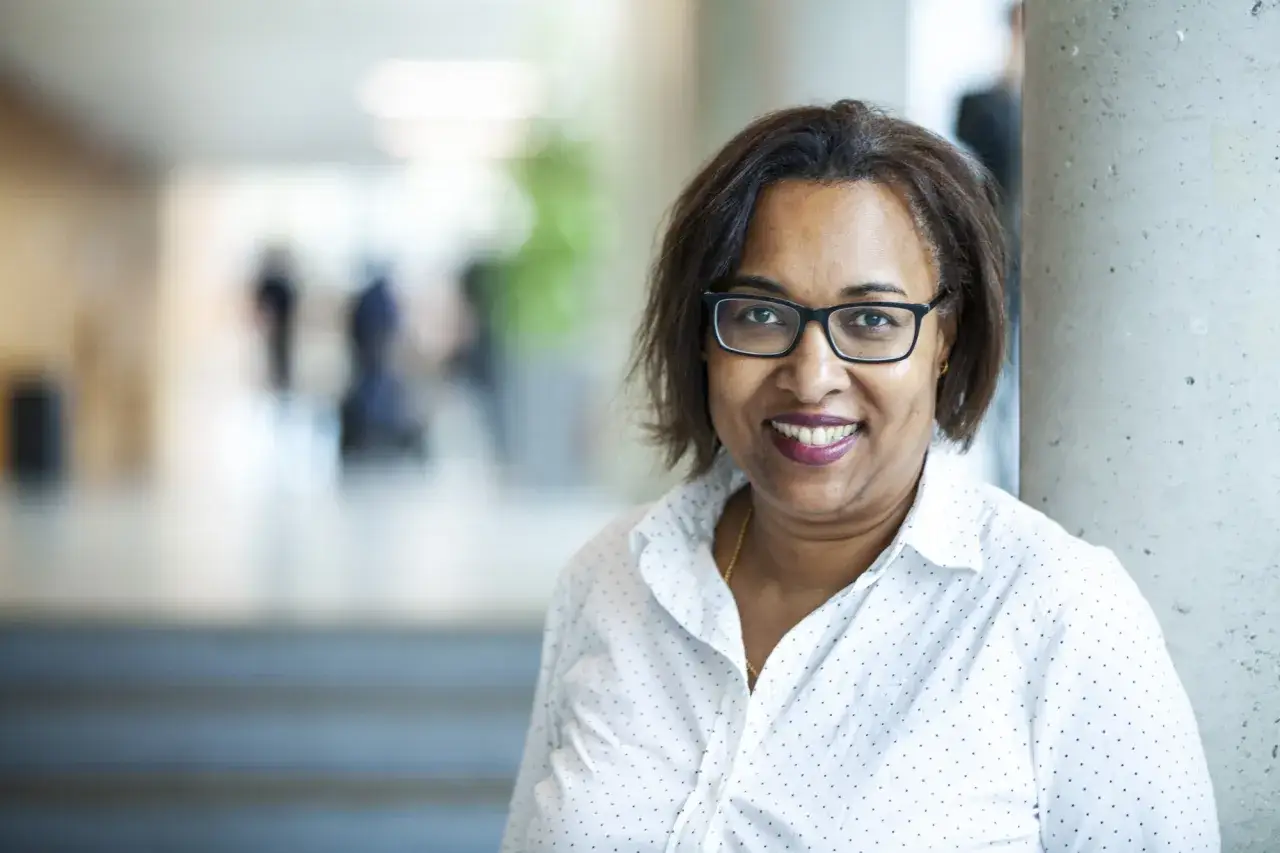Karin Geuijen, met een stralende glimlach en een witte blouse met stippen, staat in een gang.
