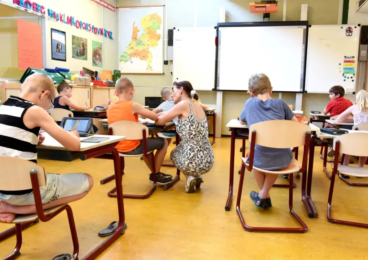 Een klas met kinderen die aan hun tafels zitten. Een lerares helpt een jongen met zijn werk. Op de muur hangt een kaart van Nederland en het alfabet.