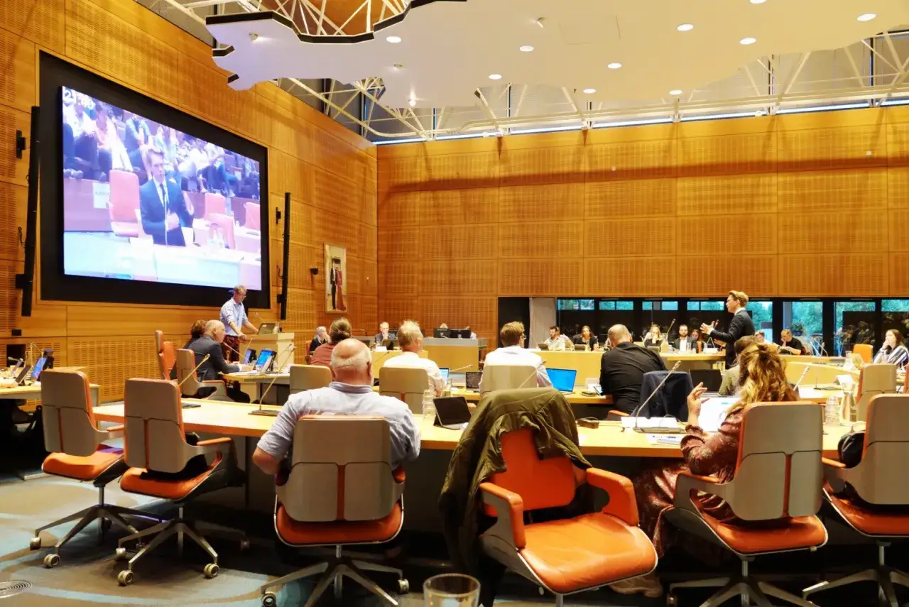 Mensen in een vergaderzaal, met een groot scherm en een spreker. Een voorbeeld van collectieve voorzieningen voor overleg.