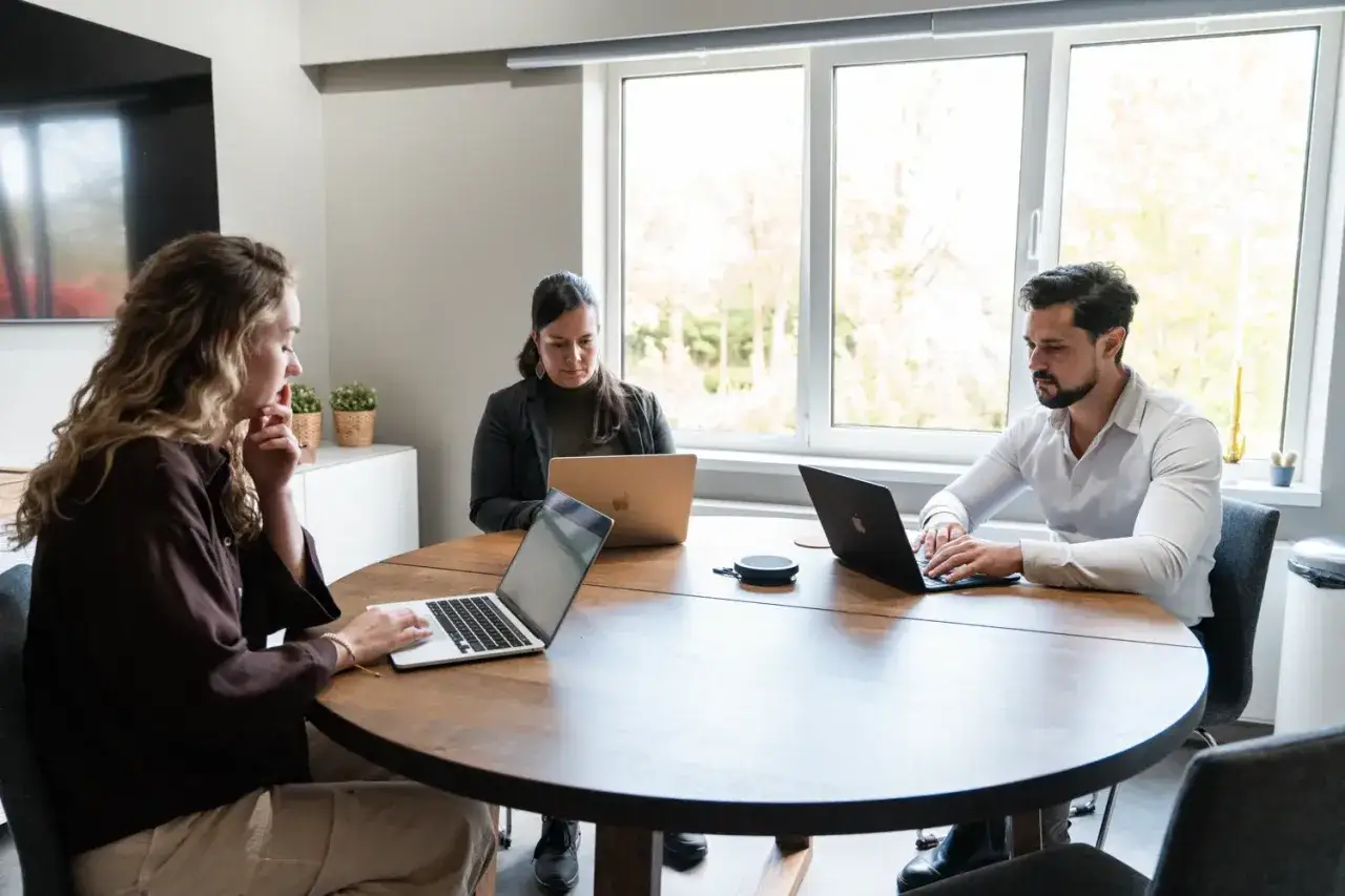 Drie collega's werken samen aan een project. Ze zitten rond een tafel met laptops. Dit is een voorbeeld van effectief leiding geven.