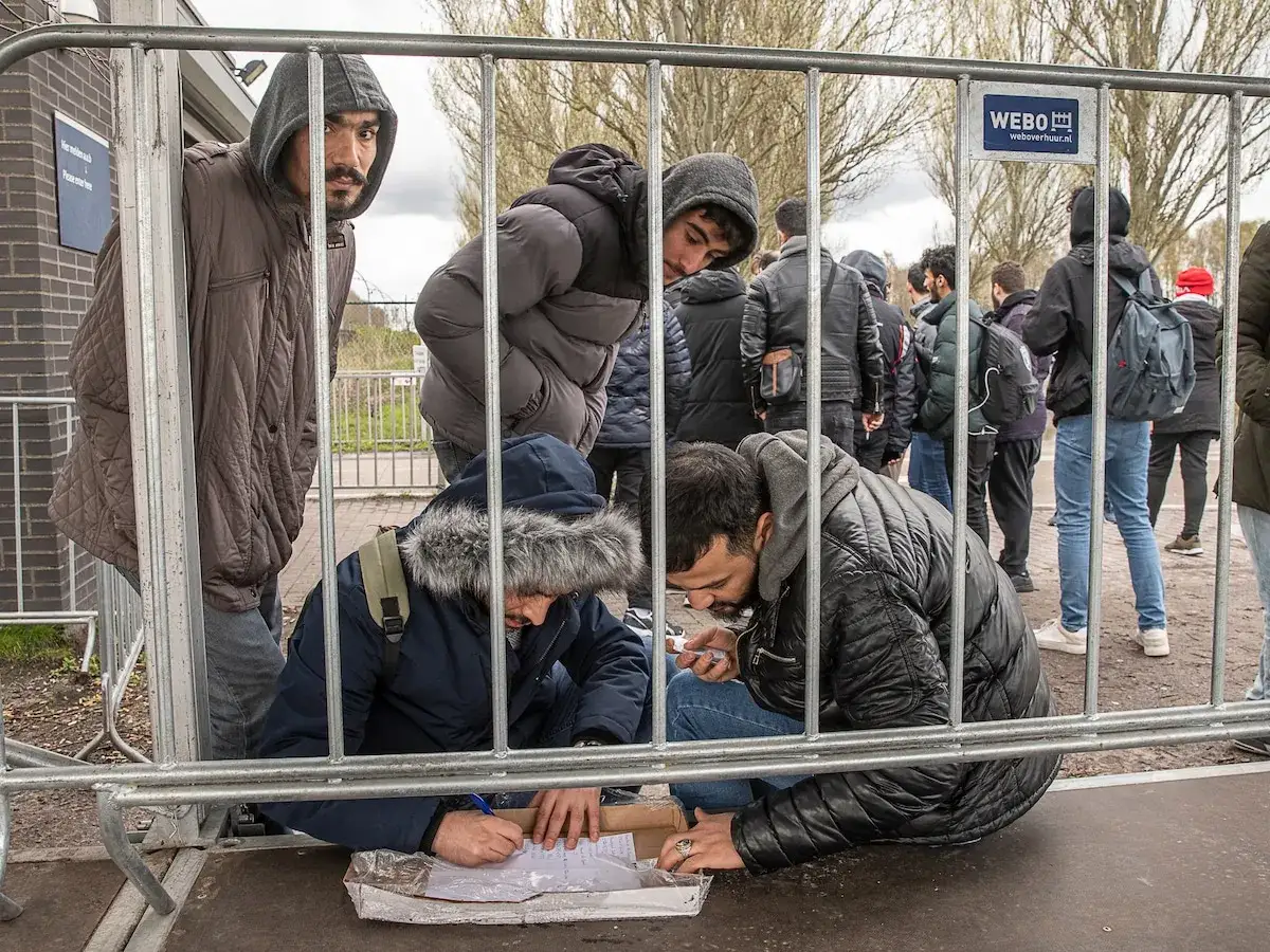 Mannen in winterkleding wachten achter hekken, sommigen schrijven papieren. Dit beeld raakt aan het vluchtelingenbeleid in Nederland.