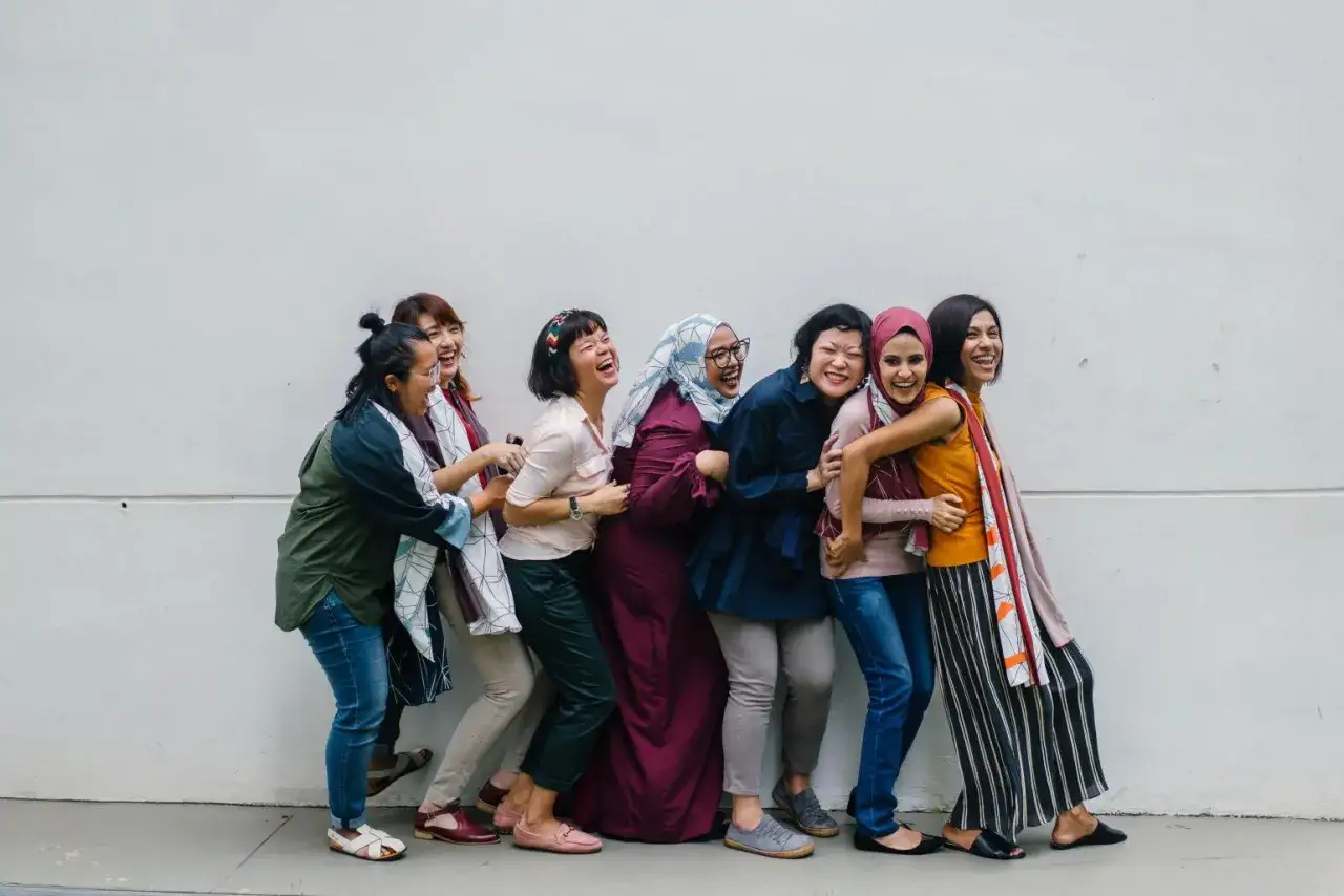 Een groep lachende vrouwen, divers in kleding en achtergrond, omhelzen elkaar. Ze vieren hun vriendschap en de kracht van hun gedeelde nationale identiteit.