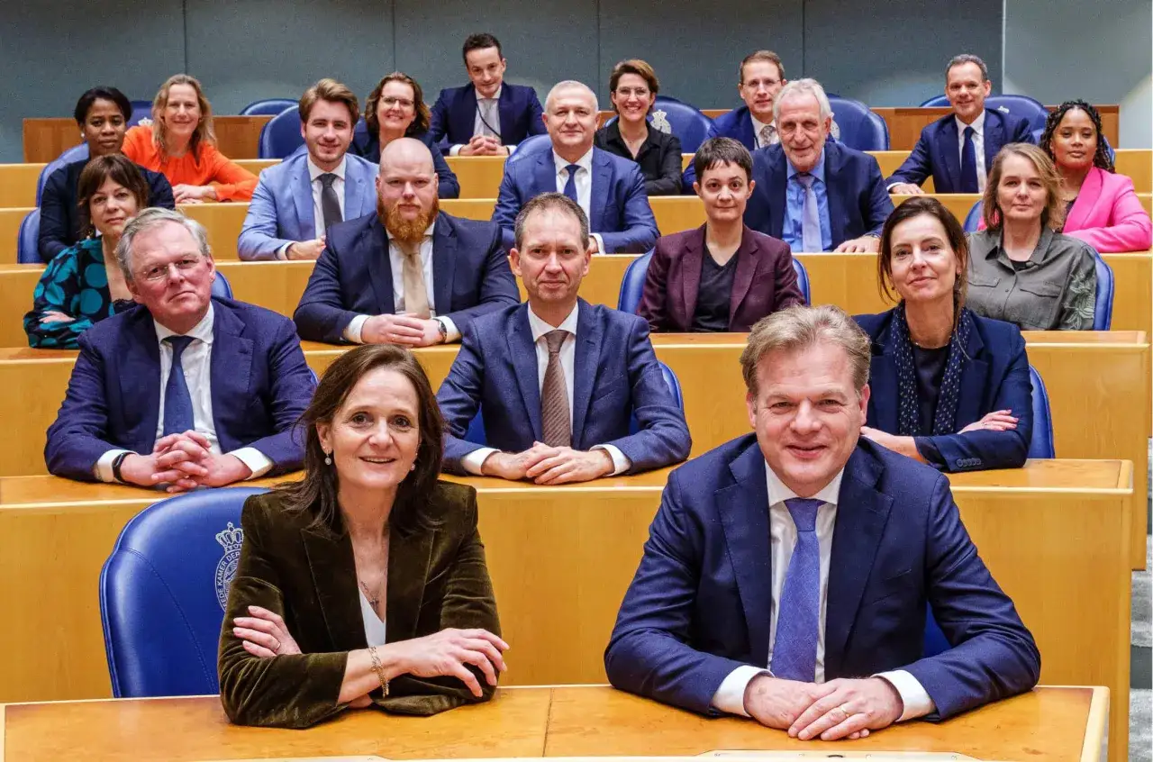 Groep politici in debat, de sociaal contract betekenis besprekend. Ze zitten in een zaal met houten tafels en blauwe stoelen.