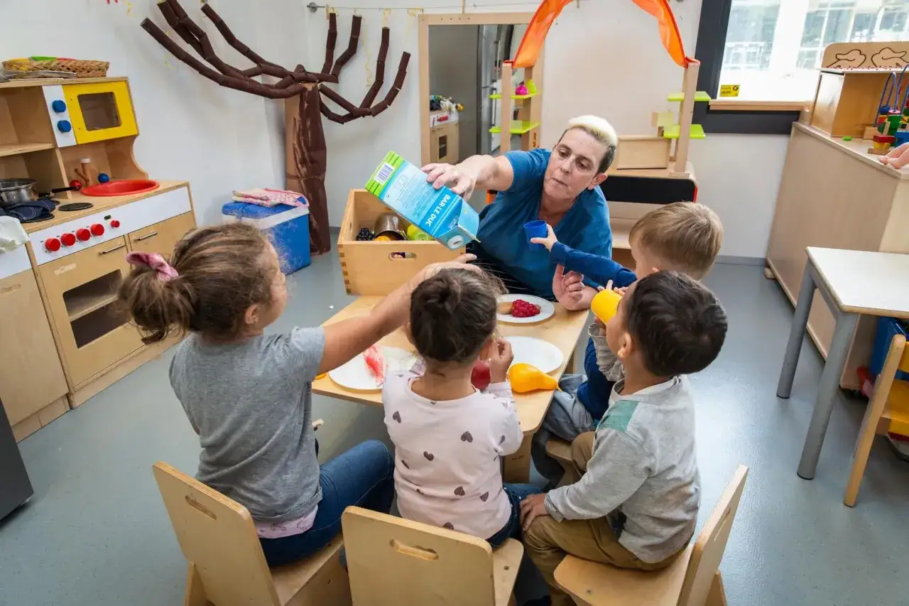 Juf schenkt sap in bekertjes, kinderen leren spelenderwijs over eten en drinken aan tafel.
