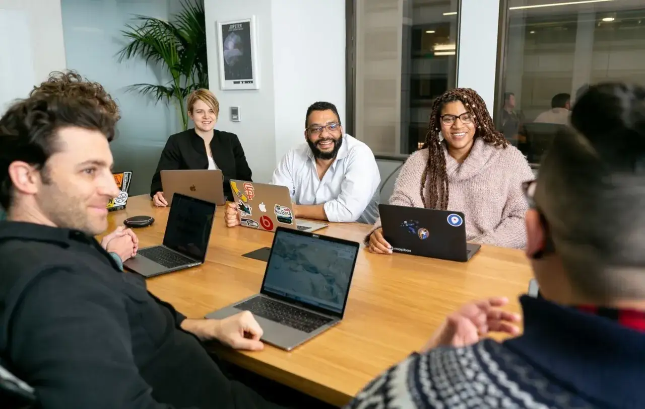Een team bespreekt idee&euml;n rond een tafel, laptops open. Dit is een voorbeeld van gedeeld leiderschap in het onderwijs, waar iedereen bijdraagt.