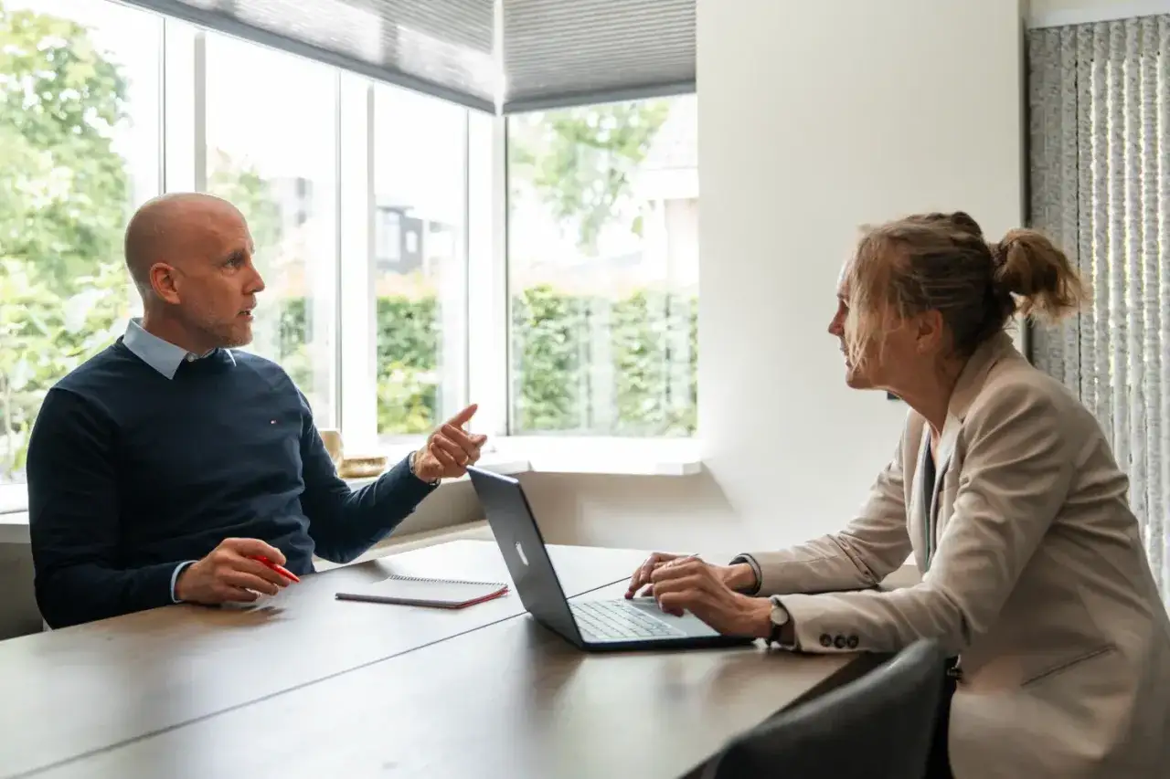 Man met rode pen probeert vrouw te overtuigen met een laptop. Ze luistert aandachtig.