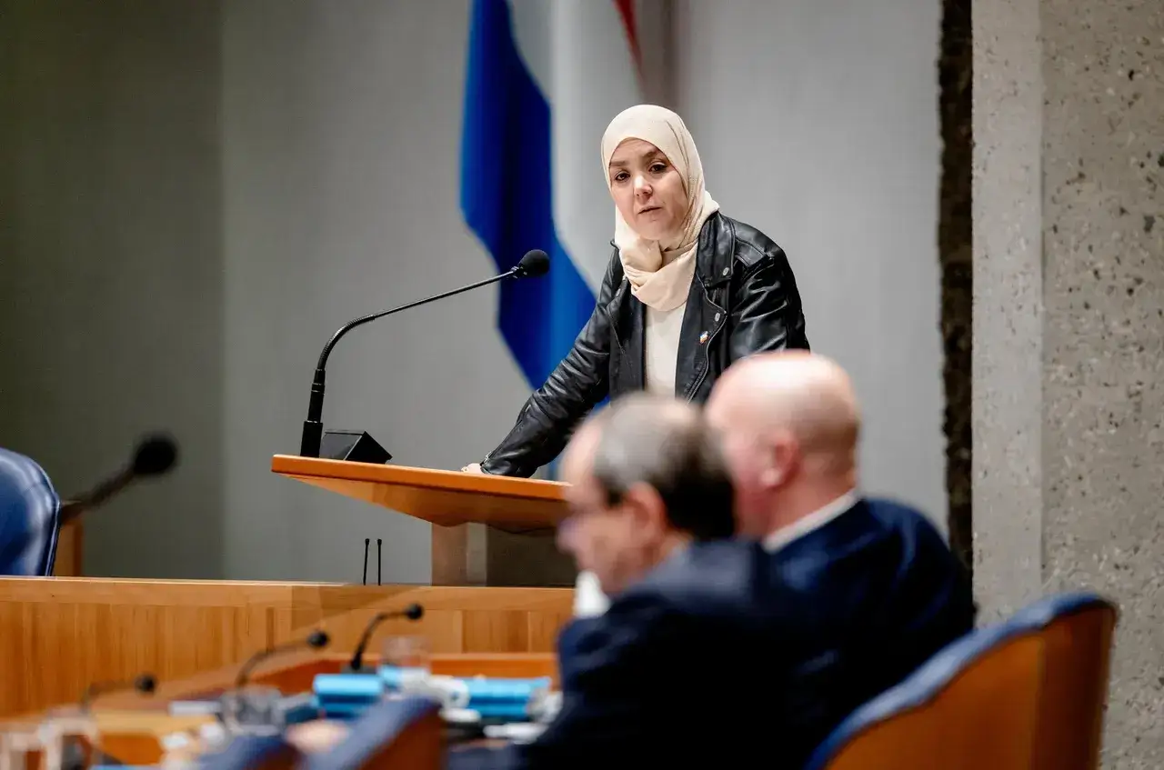 Vrouw met hoofddoek spreekt in Tweede Kamer, met GroenLinks-vlag op de achtergrond. Discussie over de Moslimbroederschap.