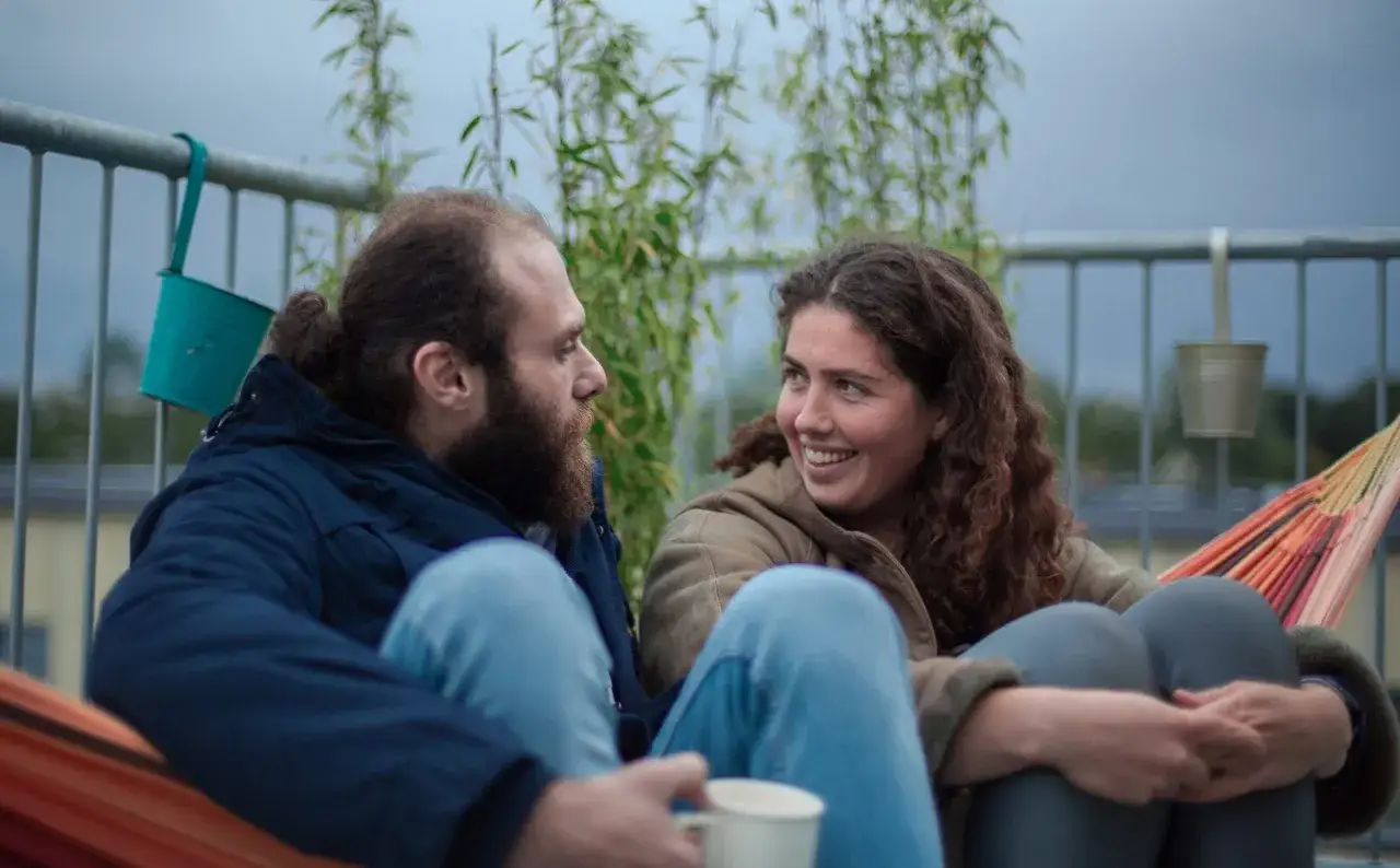 Twee syrische mensen, een man met baard en een vrouw met krullend haar, ontspannen in een hangmat en kijken elkaar lachend aan.