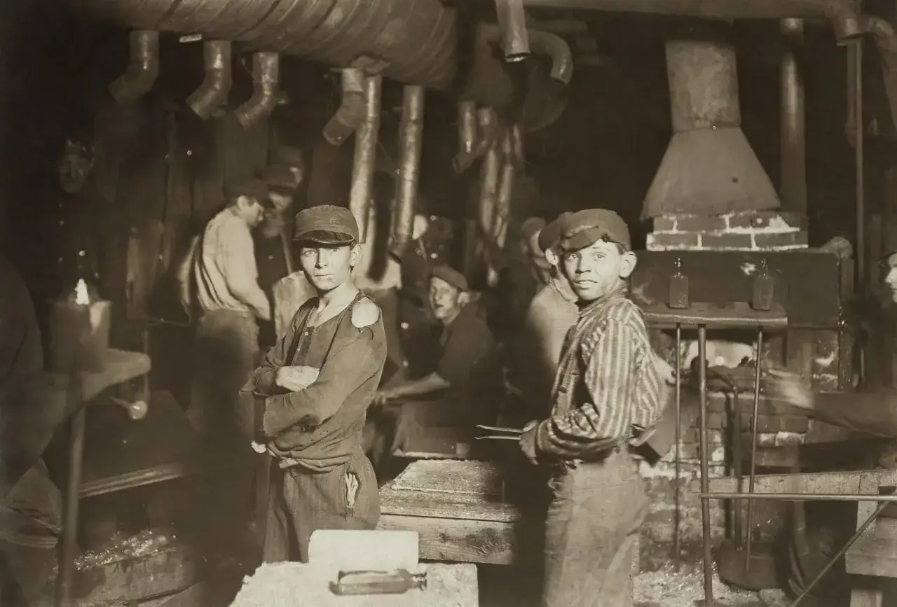 Jongens in een fabriek, een beeld dat herinnert aan het kinderwetje van Houten. Ze werken in een donkere, industriële omgeving met buizen boven hun hoofd.