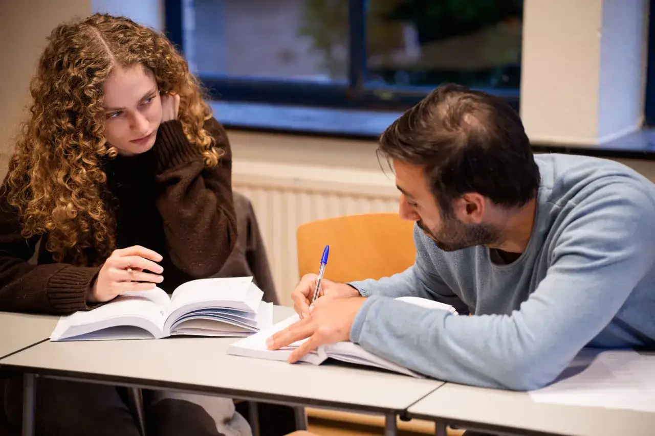 Een 23 jaar volwassen vrouw met krullend haar kijkt nadenkend naar een open boek. Naast haar schrijft een man aantekeningen.